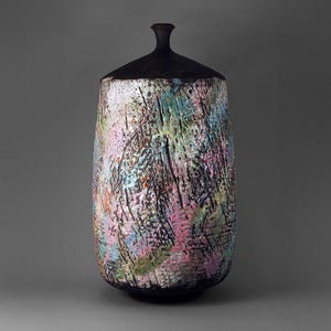 Puede incluir: Un jarrón de cerámica texturizado con tapa. El jarrón es cilíndrico con base redondeada y tapa cónica oscura. La superficie es multicolor con pinceladas rosas, azules, verdes y negras. El jarrón está sobre un fondo gris.