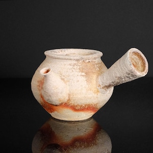 Può includere: Una teiera artigianale in ceramica bianco sporco con beccuccio e piccolo manico. La teiera ha un corpo arrotondato con una fascia marrone-rossastra vicino alla base. Il beccuccio è cilindrico e angolato.