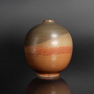 Puede incluir: Un jarrón de cerámica esférico con una abertura estrecha en la parte superior. La superficie presenta un degradado de colores, pasando de un gris carbón oscuro en la parte superior a un naranja terracota cálido y un marrón claro moteado en la base.