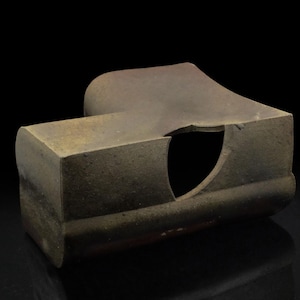 Puede incluir: Una escultura de cerámica con una superficie rugosa y texturizada. La escultura es una forma geométrica con un gran agujero en el centro. La escultura es de color marrón oscuro con toques de verde y amarillo.