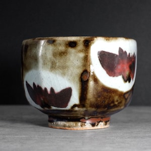 Pode incluir: Uma chávena de chá de cerâmica com formato arredondado e pé pequeno. A chávena apresenta um esmalte mosqueado castanho e branco com desenhos florais vermelho escuro. O interior é de cor creme claro e o exterior tem um aspeto texturizado.