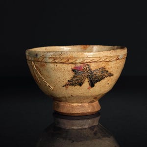 May include: A handmade ceramic bowl with a beige glaze and a decorative leaf design. The bowl features a textured rim and a small, circular base. The leaf design is black and red. The bowl is approximately 4 inches in diameter.