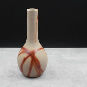 Puede incluir: Un pequeño jarrón de cerámica con cuerpo redondeado y cuello largo y delgado. El jarrón está decorado con líneas marrones, como pinceladas, sobre un fondo beige claro.