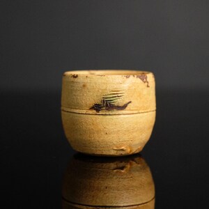 May include: A small, cylindrical, antique-style container with a light tan exterior. The container features a dark brown and green painted design. The object is set against a dark background, with its reflection visible below.