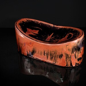 Könnte beinhalten: Eine nierenförmige Keramikschale mit glänzendem Finish. Der Innenraum ist schwarz mit rotbraunen Akzenten, während die Außenseite rot-orange mit schwarzen Streifen ist. Die Schale steht auf einer reflektierenden Oberfläche.