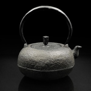 May include: A black cast iron teapot with a rounded body and a curved handle. The teapot has a textured surface and a small knob on the lid.