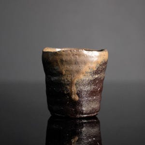 May include: A handmade ceramic cup with a rustic, textured appearance. The cup features a dark brown base that transitions to lighter brown and beige tones near the rim. The glaze has a dripped effect, adding to its unique character.