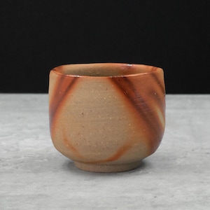 Puede incluir: Una pequeña taza de cerámica hecha a mano con base redondeada y boca ancha. La taza presenta un exterior marrón claro con patrones arremolinados de color marrón rojizo. El interior es de un tono más claro.