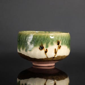 May include: A ceramic tea bowl with a textured, wavy surface. The upper portion features a green glaze that drips over a cream-coloured middle section, which is decorated with small, dark brown painted sprigs. The base is a solid dark brown.