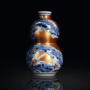 May include: A double-gourd-shaped ceramic vase with a metallic gold finish and blue and white floral designs. The vase has a narrow neck and a wide base, with a reflective surface below.