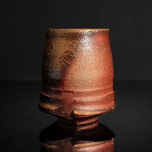Puede incluir: Una taza de cerámica rústica hecha a mano con una superficie texturizada única. La taza presenta un cuerpo cilíndrico con una base acampanada y una rica paleta de colores terrosos de marrón y rojo. La taza mide aproximadamente 10 cm de altura.