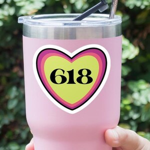 Op de afbeelding: Een roze geïsoleerde beker met een hartvormige sticker. De sticker heeft het nummer "618" in zwart op een gele achtergrond, omlijst door roze en paarse randen. Een zilveren rietje en zwarte rietjestip zitten in het deksel.