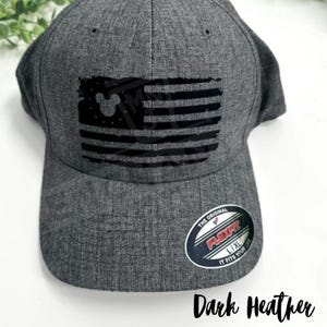 May include: Dark heather grey baseball cap with a black and white American flag design featuring a Mickey Mouse silhouette. The cap has a Flexfit label and the text "The Original Flexfit U/XL It Fits Your Life".