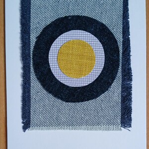 May include: A handmade greeting card with a denim fabric design. The card features a circular pattern with a yellow centre, surrounded by a navy blue ring, and a light grey and white chequered pattern. The denim fabric has frayed edges.