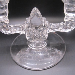 Può includere: Portacandele in vetro trasparente a due bracci. Il portacandele ha una base rotonda e dettagli ornati. Le braccia si incurvano verso l'alto per sostenere le candele. Il vetro è trasparente.