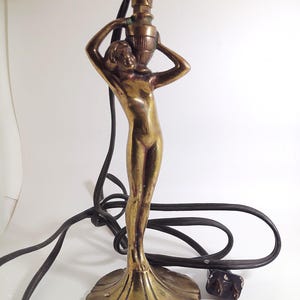 Pode incluir: Um candeeiro de mesa em latão com a figura esculpida de uma pessoa sobre uma base circular canelada, segurando um vaso acima da cabeça. O candeeiro inclui um casquilho de metal com um interruptor rotativo preto e um cabo elétrico preto.
