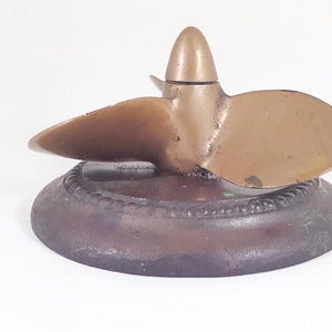 May include: A metallic boat propeller with three curved blades and a central conical hub, mounted on a circular, dark-toned base featuring a decorative beaded edge. The object displays a weathered, aged finish with visible patina.