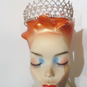 May include: A silver-toned tiara with clear rhinestones sits atop a mannequin head. The tiara features an openwork design with a series of sparkling stones. The mannequin has closed eyes with long eyelashes, red lips, and orange hair.