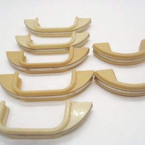 May include: A collection of vintage, cream-coloured cabinet handles. Each handle has a curved design with a central, recessed area and a thin, silver-coloured accent stripe. The handles are arranged on a white surface.