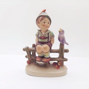 May include: A ceramic figurine of a young boy sitting on a wooden fence, with a purple bird perched beside him. The boy wears a hat, green shirt, and brown overalls. The figurine is on a round base.