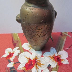 May include: A small, antique brass vase with an ornate design. The vase has a rounded body and a narrow neck. It is decorated with a floral pattern and has two small handles on the sides.