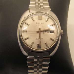 Puede incluir: Un reloj de pulsera automático Burien JJ vintage con una correa y caja de metal plateado. La esfera del reloj es de color claro y muestra el día y la fecha. El reloj está sobre una superficie negra.