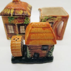 May include: A set of three ceramic salt and pepper shakers shaped like cottages with thatched roofs. The shakers are decorated with brown, green and yellow paint. The salt shaker is a windmill with a brown wheel. The pepper shaker is a cottage with a brown roof and a small window. The sugar shaker is a cottage with a brown roof and a small window.