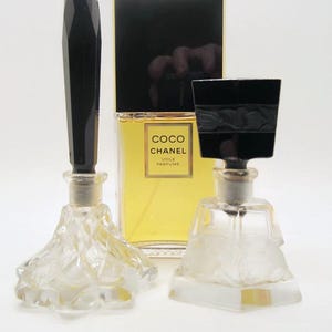 May include: Three perfume bottles and a perfume box. The perfume bottles have clear glass bases and black stoppers. The perfume box is black and gold with the words "COCO CHANEL VOILE PARFUME" on the front.
