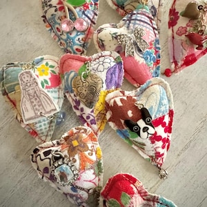 May include: A collection of handmade fabric heart ornaments in various colors and patterns. Each heart features unique embellishments, including charms, beads, and appliqués of a cat, dog, and other designs. The hearts are arranged on a light-colored surface.