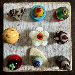 May include: A collection of nine decorative knobs and pulls in various shapes, colors, and materials, arranged on a white-painted wooden board. The knobs include floral, geometric, and abstract designs, with accents of red, yellow, green, and blue.