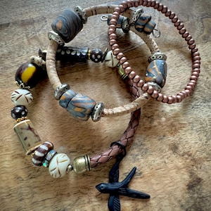 May include: Three handcrafted bracelets with unique beads and charms. One bracelet features a black bird charm. The beads are in various colors, including brown, tan, and blue. The bracelets are on a wooden surface.