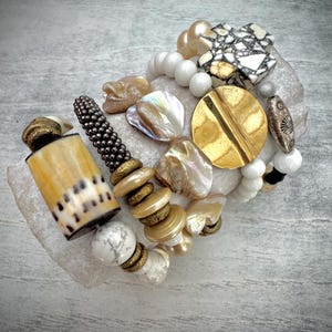 May include: A stack of bracelets with white, gold, and brown beads. The bracelets feature a variety of materials, including shell, wood, and metal.