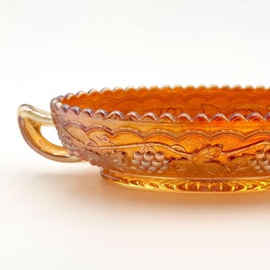 May include: An iridescent orange glass dish with a scalloped rim and two handles. The oval-shaped bowl features a grape and leaf design. This decorative piece is likely vintage and suitable for serving.