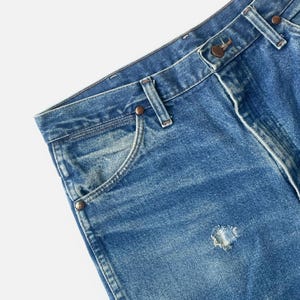 Puede incluir: Primer plano de unos vaqueros de mezclilla azules con un agujero desgastado. Los vaqueros tienen un dise&ntilde;o cl&aacute;sico de cinco bolsillos, trabillas y cierre de bot&oacute;n y cremallera. La mezclilla tiene un aspecto descolorido y desgastado.