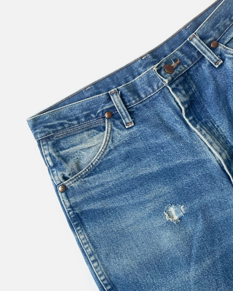 Puede incluir: Primer plano de unos vaqueros de mezclilla azules con un agujero desgastado. Los vaqueros tienen un dise&ntilde;o cl&aacute;sico de cinco bolsillos, trabillas y cierre de bot&oacute;n y cremallera. La mezclilla tiene un aspecto descolorido y desgastado.