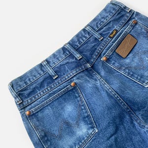 May include: Close-up of the back of blue denim jeans. The jeans feature a brown leather patch with the "Wrangler" logo and copper rivets. The back pockets have a signature "W" stitching design.