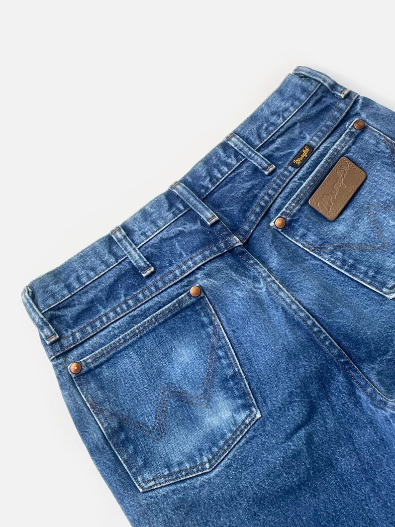 May include: Close-up of the back of blue denim jeans. The jeans feature a brown leather patch with the "Wrangler" logo and copper rivets. The back pockets have a signature "W" stitching design.