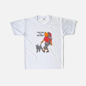 K&ouml;nnte beinhalten: Wei&szlig;es T-Shirt mit einer Cartoon-Illustration von Tim und Struppi. Tim tr&auml;gt ein rotes Hemd, eine graue Hose und einen Rucksack. Der Text "TINTIN IN VIETNAM" ist &uuml;ber der Figur aufgedruckt. Der Hund ist grau.