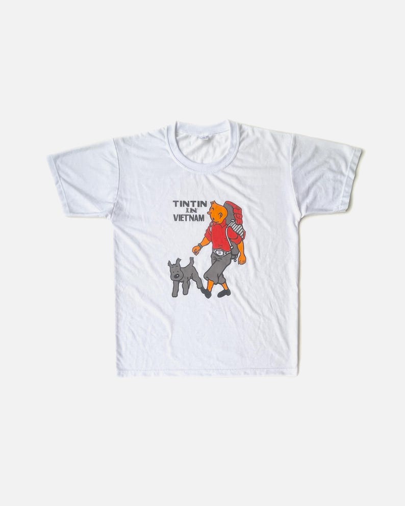 K&ouml;nnte beinhalten: Wei&szlig;es T-Shirt mit einer Cartoon-Illustration von Tim und Struppi. Tim tr&auml;gt ein rotes Hemd, eine graue Hose und einen Rucksack. Der Text "TINTIN IN VIETNAM" ist &uuml;ber der Figur aufgedruckt. Der Hund ist grau.