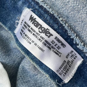 Puede incluir: Primer plano de la etiqueta de unos vaqueros Wrangler azules. La etiqueta blanca tiene texto negro, incluyendo la marca, la talla 32 x 32 y las instrucciones de lavado. Los vaqueros son de 100% algod&oacute;n.