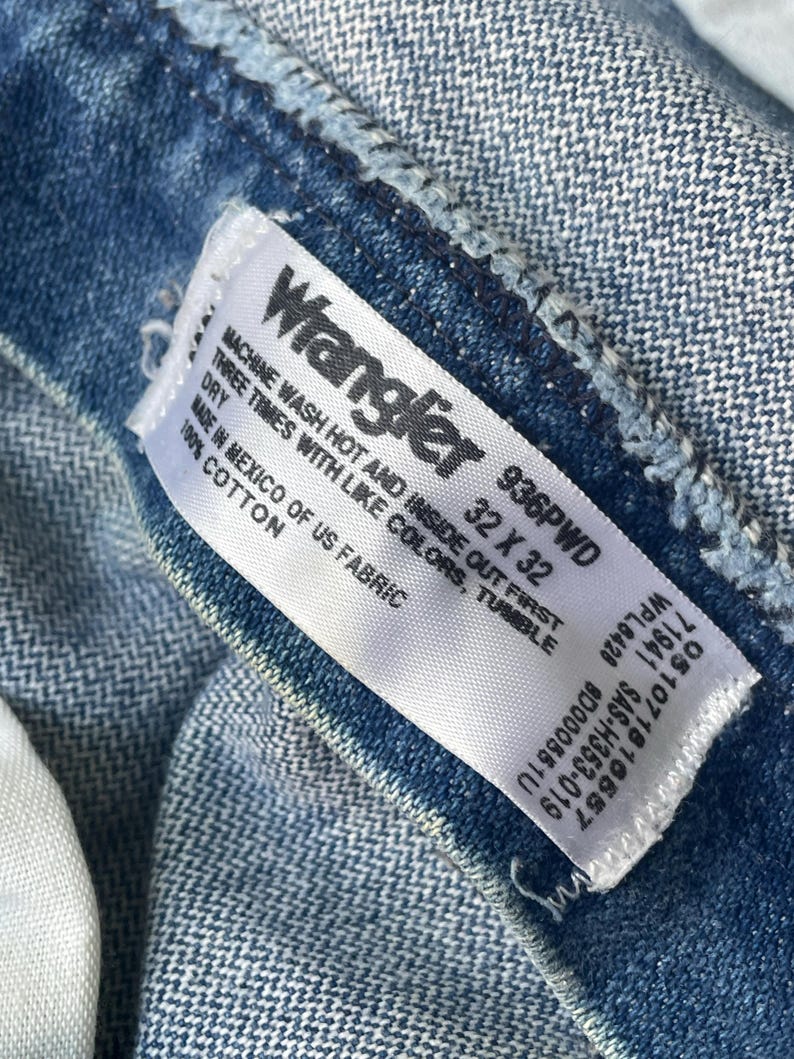 Puede incluir: Primer plano de la etiqueta de unos vaqueros Wrangler azules. La etiqueta blanca tiene texto negro, incluyendo la marca, la talla 32 x 32 y las instrucciones de lavado. Los vaqueros son de 100% algod&oacute;n.