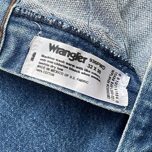 May include: A close-up shot of a blue denim garment with a white label. The label reads "Wrangler" and includes size information: 32 x 30 inches (81 x 76 cm). The label also provides washing instructions and fabric details. The garment is made of 100% cotton.