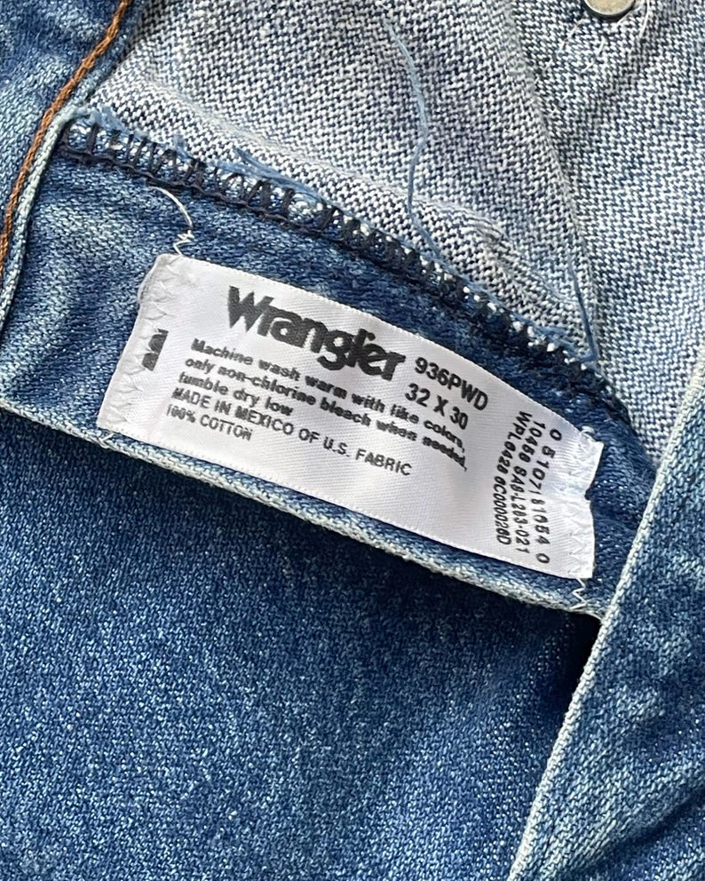 May include: A close-up shot of a blue denim garment with a white label. The label reads "Wrangler" and includes size information: 32 x 30 inches (81 x 76 cm). The label also provides washing instructions and fabric details. The garment is made of 100% cotton.