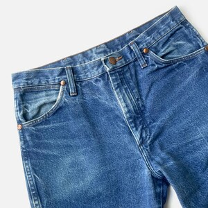May include: Close-up of blue denim jeans. The jeans feature a classic five-pocket design with copper-coloured rivets and a button closure. The denim has a faded appearance, with lighter areas on the front and back.