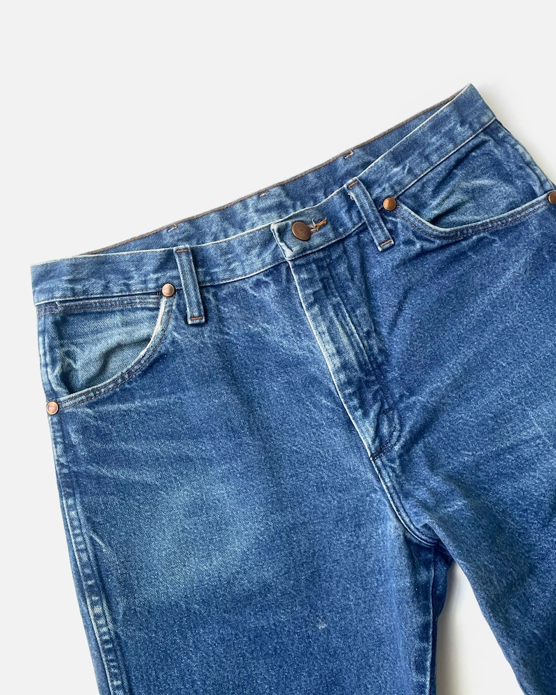 May include: Close-up of blue denim jeans. The jeans feature a classic five-pocket design with copper-coloured rivets and a button closure. The denim has a faded appearance, with lighter areas on the front and back.
