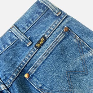 Puede incluir: Primer plano de unos vaqueros azules con el logotipo de Wrangler en una peque&ntilde;a etiqueta negra. Los vaqueros tienen un dise&ntilde;o cl&aacute;sico de cinco bolsillos con remaches cobrizos y costuras detalladas. La mezclilla tiene un aspecto descolorido.