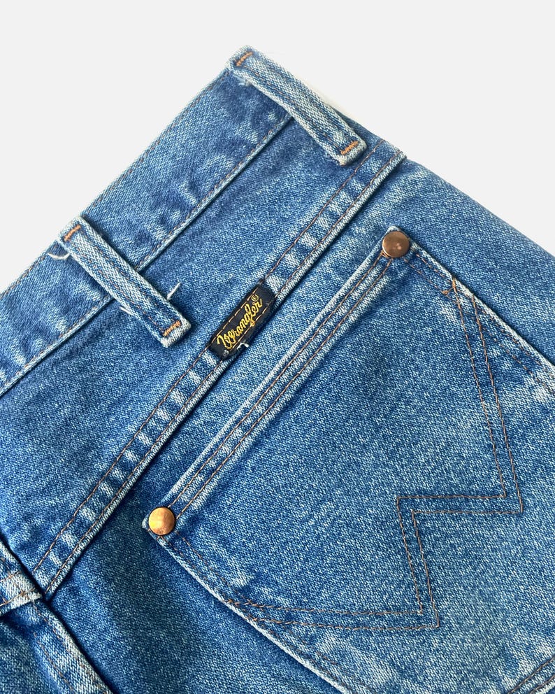 Puede incluir: Primer plano de unos vaqueros azules con el logotipo de Wrangler en una peque&ntilde;a etiqueta negra. Los vaqueros tienen un dise&ntilde;o cl&aacute;sico de cinco bolsillos con remaches cobrizos y costuras detalladas. La mezclilla tiene un aspecto descolorido.