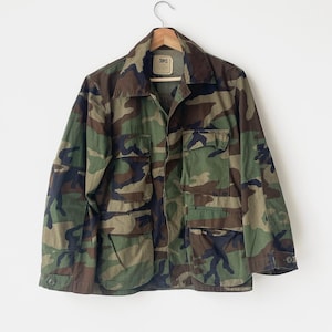 Könnte beinhalten: Tarnmusterjacke mit mehreren Taschen und einem Knopfverschluss. Die Jacke hängt an einem Holzbügel. Die Farbpalette umfasst Grüntöne, Braun und Schwarz.