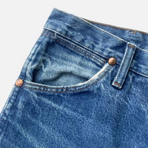 May include: Close-up of blue denim jeans pocket with copper rivets and brown stitching. The jeans have a classic five-pocket design and a faded wash. The pocket is slightly worn, adding to the vintage aesthetic.