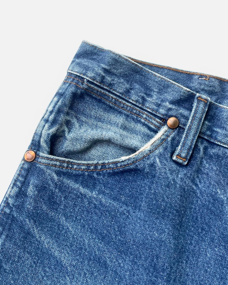 May include: Close-up of blue denim jeans pocket with copper rivets and brown stitching. The jeans have a classic five-pocket design and a faded wash. The pocket is slightly worn, adding to the vintage aesthetic.